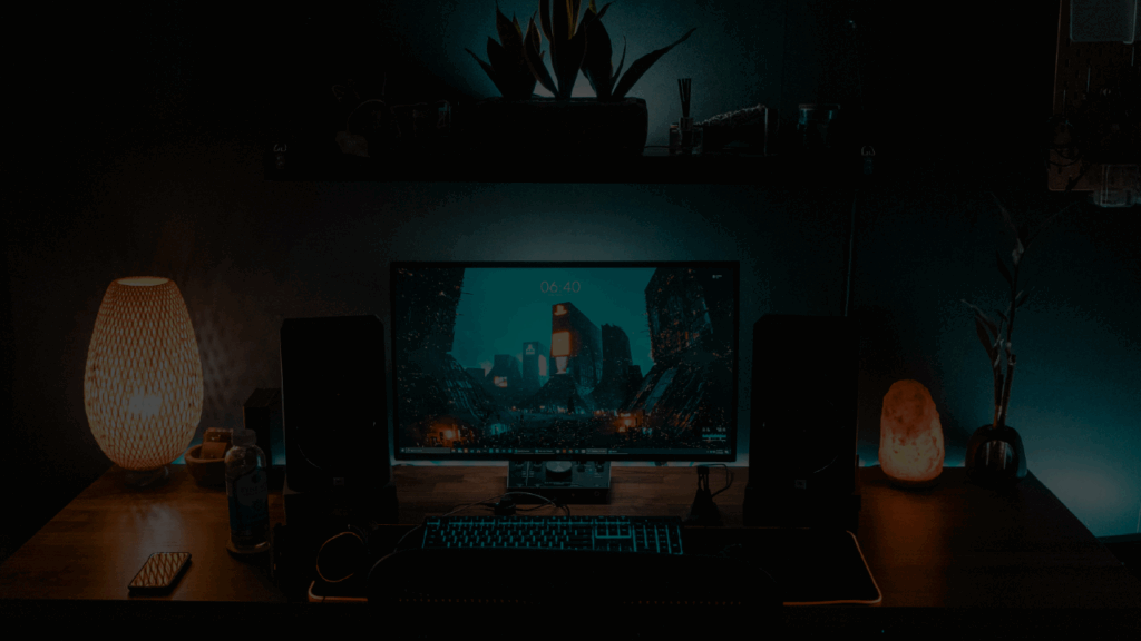 Desk on which a PC monitor and backlit keyboard sit in dimly lit conditions. There are decorative elements such as lamps and plants. Behind the desk is an LED strip that runs the full width of the desk. Representing the AI in the future of gaming