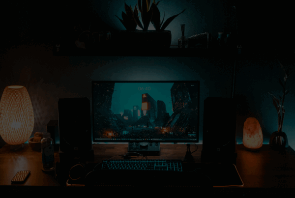 Desk on which a PC monitor and backlit keyboard sit in dimly lit conditions. There are decorative elements such as lamps and plants. Behind the desk is an LED strip that runs the full width of the desk. Representing the AI in the future of gaming
