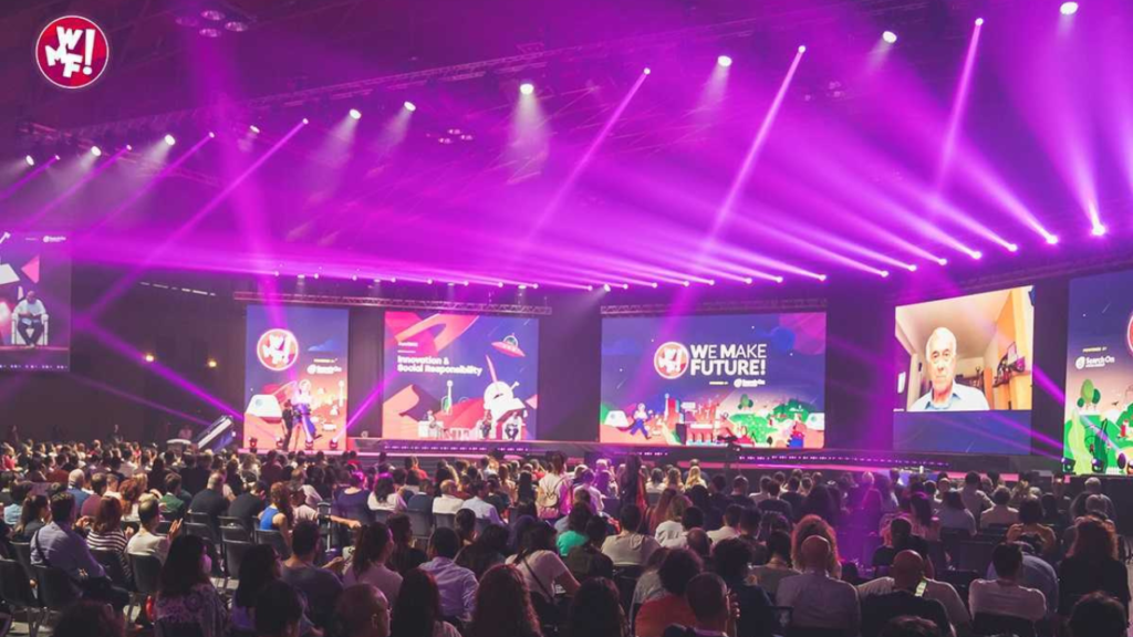 Una grande conferenza con un pubblico seduto, illuminazione colorata sul palco, schermi multipli con la scritta “We Make Future” e un relatore in videochiamata.