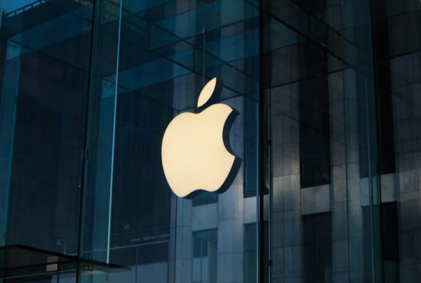Logo Apple illuminato su una vetrina di un Apple Store moderno con riflessi di edifici in vetro sullo sfondo.